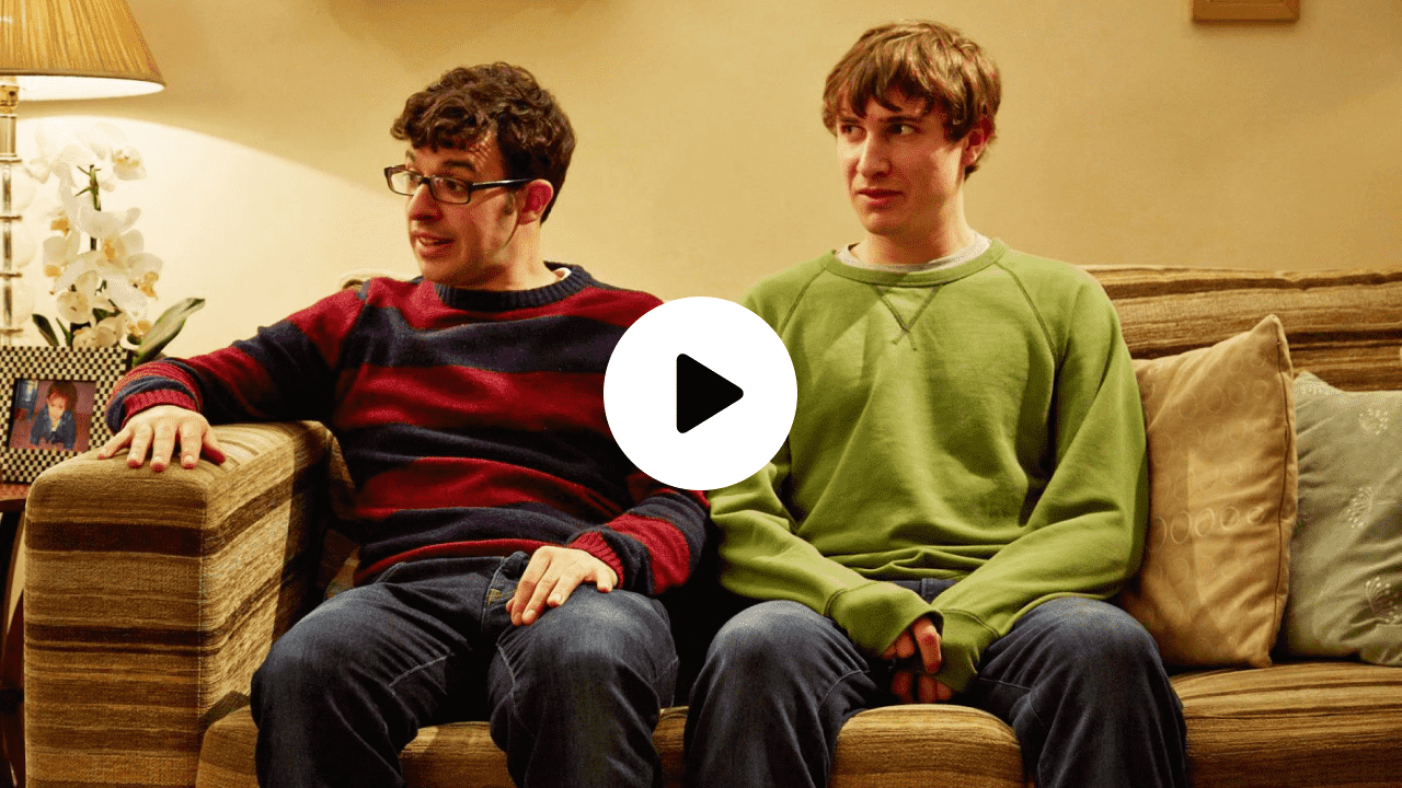 Scene from the British sitcom Friday Night Dinner, showing two main characters sitting awkwardly on a sofa, capturing the show’s quirky humor and family-centered chaos.
